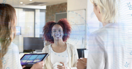 Smiling woman in knitted cardigan holding coffee cup and talking in modern office, tablet visible....