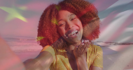 Laughing woman wearing yellow dotted blouse blowing kiss on beach, holding phone with flag graphic....