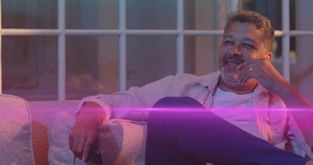 Smiling man wearing button-up over tee, leaning back on sofa at home, holding remote, showing ring....