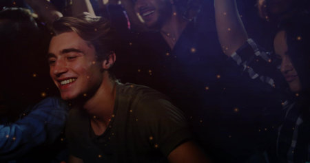 Smiling adult male leaning forward in olive-green T-shirt at nightclub, warm lights and raised arms....