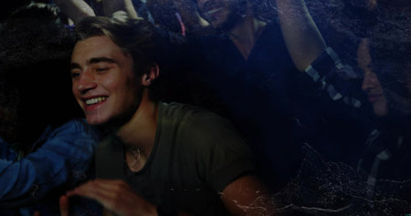Smiling male in olive T-shirt leaning forward in crowded dim club, necklace visible, raised arms....