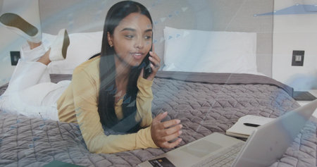 Lying adult woman wearing pale yellow top, white pants holding smartphone, looking at laptop on bed....