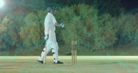 Walking batsman carrying wooden bat wearing white kit pads near stumps on night pitch, copy space....