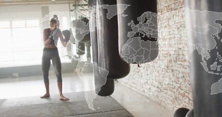 Training female boxer in sports bra and capris, wearing boxing gloves, striking heavy bag in studio....