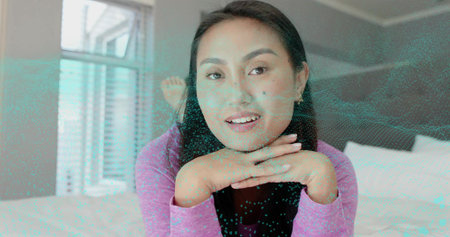 Leaning forward Asian woman wearing pink-lilac top, chin on hands in bedroom with cyan hologram....