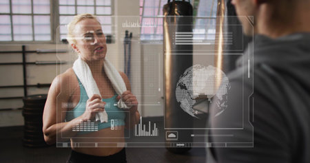Talking woman in turquoise sports bra pausing at gym with towel, punching bag, holographic data....