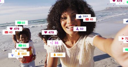 Smiling woman in crochet top holding smartphone for selfie on wet sand, with floating notifications....