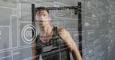 Studying man in black sleeveless tank examining data on transparent panel at studio with HUD. Glass,...