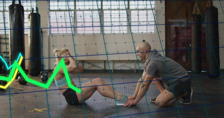 Doing crunches, trainee in sports kit on rubber mat in gym, trainer holding feet, neon graph....