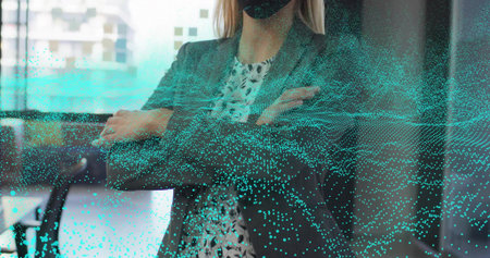 Standing woman wearing dark blazer and printed blouse masked crossing arms in lobby with teal data....