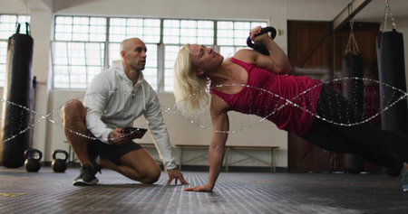Performing client in pink tank top holding kettlebell in side plank at studio, trainer with tablet....