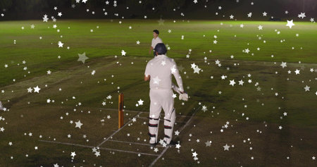 Batting helmeted batsman in white kit at crease by wooden stumps on floodlit pitch, star overlays. Cricket, nighttime, floodlights, grass, field, bat, duoの写真素材