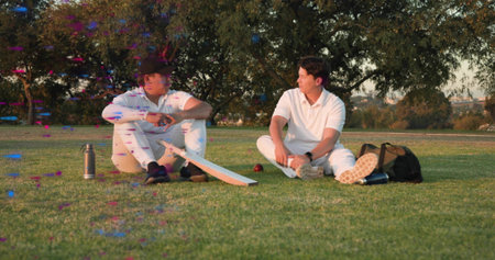 Sitting cricket teammates wearing white kit, chatting on park field with cricket bat and red ball....
