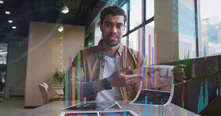 Presenting man wearing brown jacket gesturing at printed charts with translucent overlays in office....