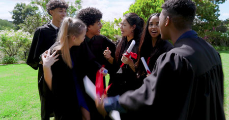 Celebrating graduates in black gowns hugging on college lawn, holding diplomas with red ribbons....