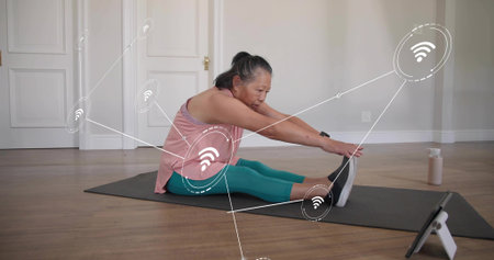 Stretching Chinese woman in pink top reaching feet on home mat with tablet and Wiâ€‘Fi...
