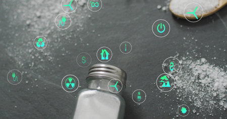 Showing metal-capped glass shaker spilling salt on dark slate, wooden spoon and green icons. Macro,...