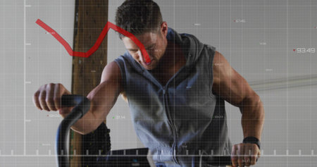 Pedaling mid adult man gripping bike handlebars in workout studio, sleeveless hoodie and smartwatch....