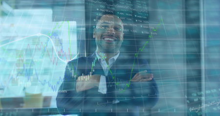 Standing trader wearing dark suit crossing arms smiling in modern office at monitors with charts....