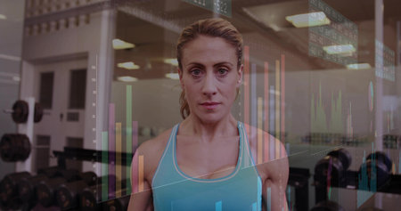 Standing mid-adult woman in light blue tank top in gym weight area, showing dumbbells, HUD data....