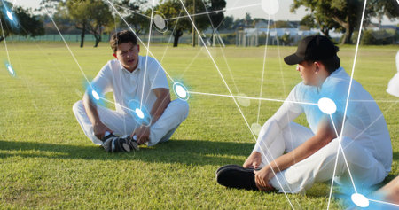 Sitting teammates in white kit, black cap, adjusting shoes and talking on field, digital overlay....