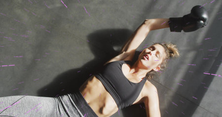Lying athletic woman wearing black sports bra, gray leggings, holding boxing glove on gym mat....