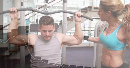 Pulling man with sleeveless grey top performing lat pulldown at gym, trainer guiding curved bar....