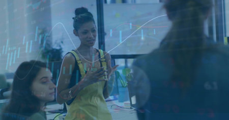 Leading woman in yellow dress gesturing and speaking at office meeting with laptops, stock charts....