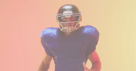 Facing male football player wearing dark helmet with facemask and blue shoulder pads in studio. Headgear, jersey, gridiron, torso, navy, gradient, highkeyの写真素材
