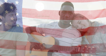 Sitting trio leaning, holding acoustic guitar on shore in casual clothes, American flag overlay....