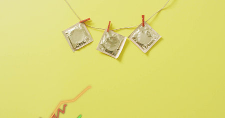 Hanging three silver foil condom packets on twine over pale yellow backdrop, neon wires lower-left....