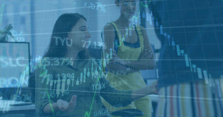 Gesturing seated woman explaining charts at desk with monitors, standing yellow-top woman, overlay....