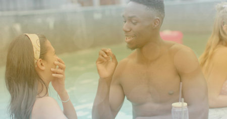 Smiling shirtless man leaning toward woman with printed headband and bracelet by pool rim with cup....