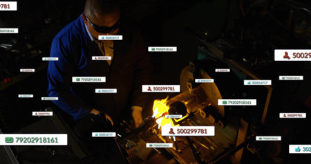 Using torch, man in blue jacket, goggles and gloves heating metal on workshop bench with tags....