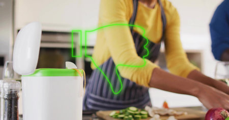 Chopping woman in yellow top, apron slicing cucumbers on kitchen counter, near green-rim trash can. Homecook, teamwork, vegetables, cuttingboard, mealprep, welllit, brightの写真素材