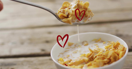 Lifting spoon dripping milk and cornflakes over white bowl on wooden table, two red heart doodles....