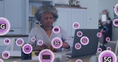 Reviewing senior woman in embroidered blouse sorting papers, using laptop at kitchen table with 5G....