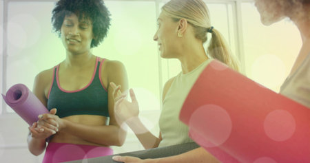 Standing three women wearing athletic clothing and holding rolled yoga mats by large studio windows....