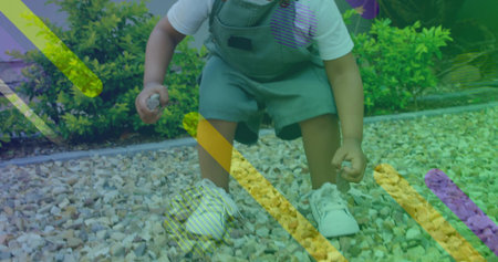 Squatting toddler holding small stones in gravel bed, wearing green overalls and white sneakers....