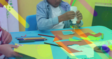 Squeezing boy in denim shirt applying white glue onto paper shapes at turquoise table, with pencils....