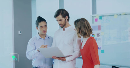 Discussing team wearing business attire leaning over silver laptop at office with glass whiteboard....