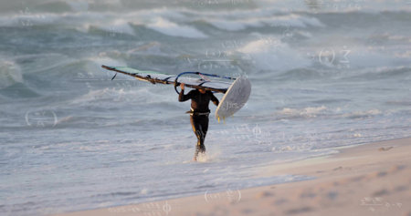 Vertical video: Leaving surf man in wetsuit holding board and sail above at beach, wearing harness....
