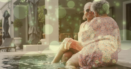 Chatting two senior women dipping feet in pool water at patio, wearing sunglasses and print dress....