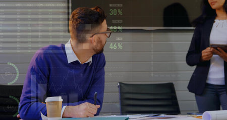 Leaning seated man in blue sweater holding pen amid papers, coffee, tablet and monitor, copy space....