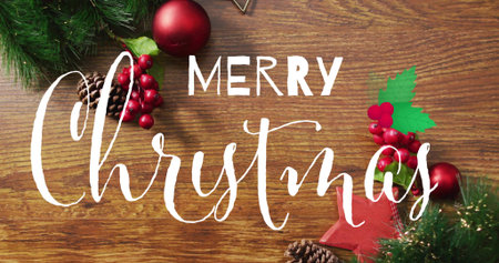 Showing centered white script Merry Christmas on wood tabletop, with evergreen sprigs, red baubles....