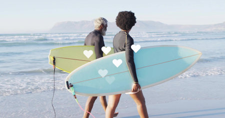 Walking two surfers toward ocean on beach, carrying surfboards with leashes, wearing black wetsuits....