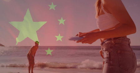 Holding round disc, woman wearing white crop top, denim jeans standing at shoreline, flag overlay....