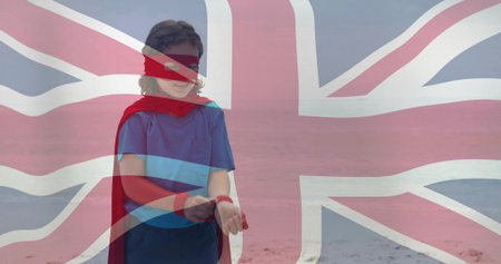 Boy in red cape adjusting wristbands on beach, wearing mask and blue T-shirt, Union Jack overlay. Child, superhero, mantle, blindfold, flag, shore, sandの写真素材
