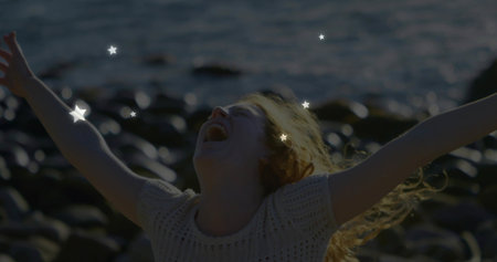 Laughing woman in knit top raising arms on pebble shore with star overlays, sun-backlit hair, sea....