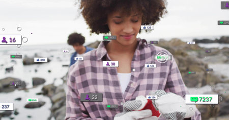 Handling small red object, woman in purple-plaid shirt and gloves on rocky shore with social badges....